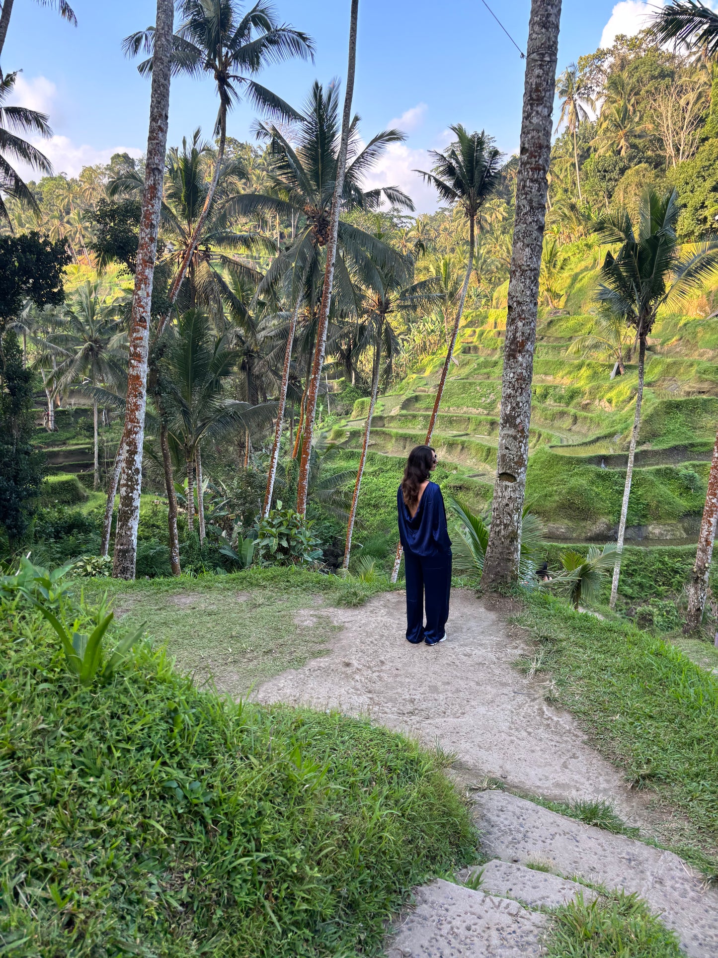 Person standing on a path surrounded by palm trees and lush greenery, Bali, Indonesia. Person wearing a satin silk set in dark blue, cowl-draped back side. RUH Draped cowl-back satin silk top from RUH Aurora Set, slow fashion luxury with cultural fusion aesthetics. Low-rise wide-leg satin silk pants from RUH Aurora Set, handmade by artisans in India with premium finish.