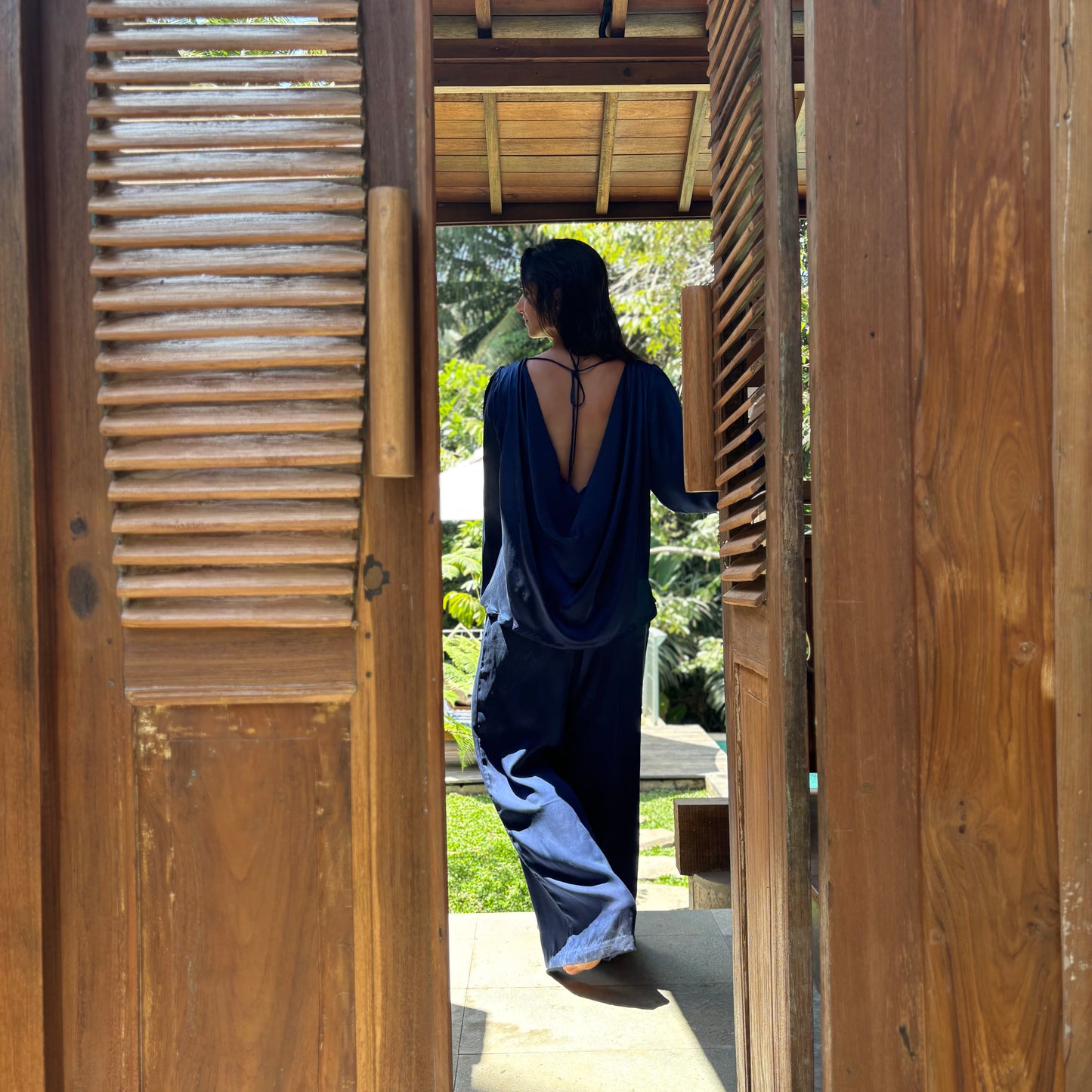 woman standing in door opening. wearing all satin silk dark blue set. backside shirt has a cowl draped back side. easygoing elegance relaxed fit set.