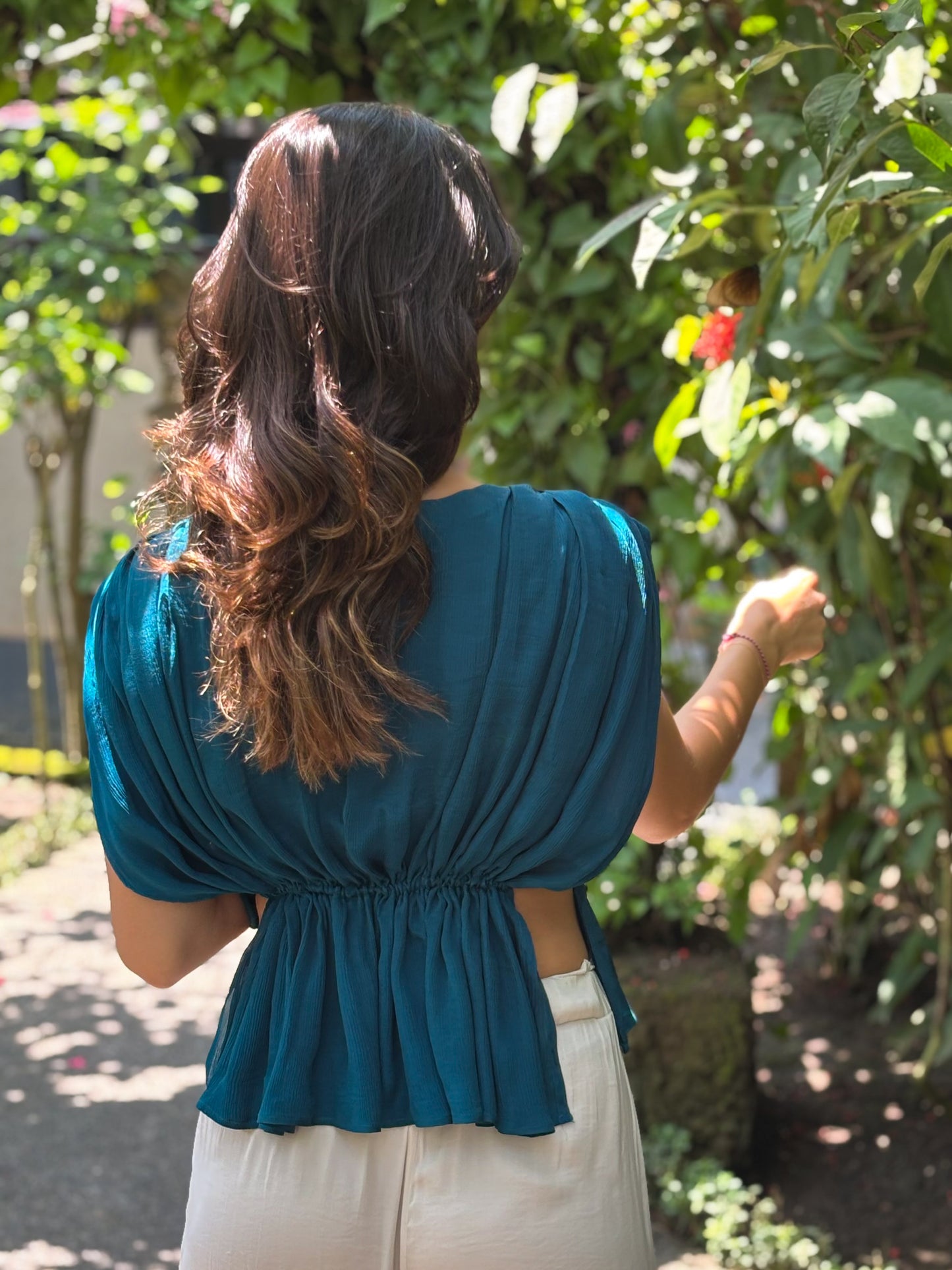 Woman wearing a teal top with a blurred green outdoor background. Silk chiffon top in turquoise with a low neck and adjustable tie around waist.