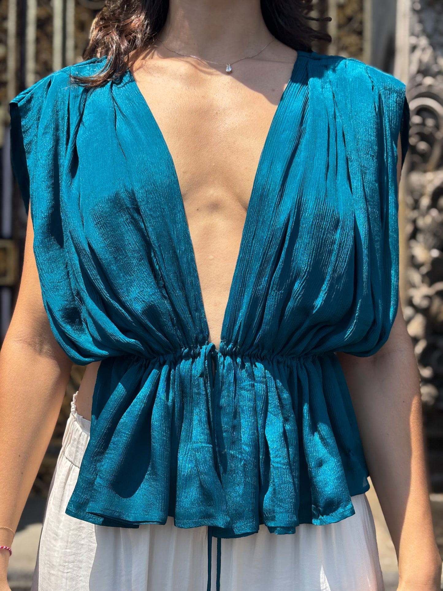 Teal sleeveless top with open back worn by a person, with a blurred background. Silk chiffon top in turquoise/teal with a low neck and adjustable tie around waist.