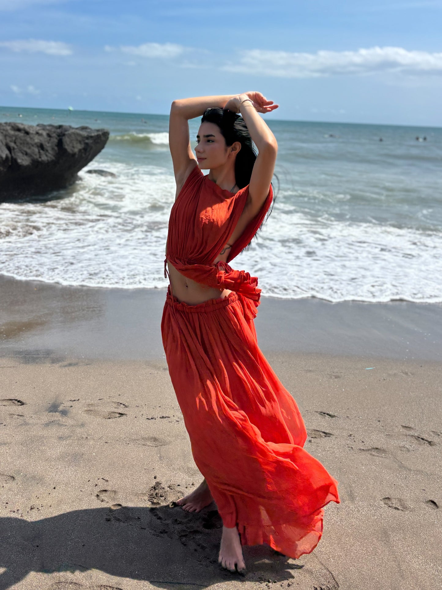 Woman in a red dress standing on a beach with ocean and sky in the background. RUH Agni Silk Set in orange rust – luxury slow fashion coord crafted by artisans in Chandigarh, blending Indian heritage with modern design.