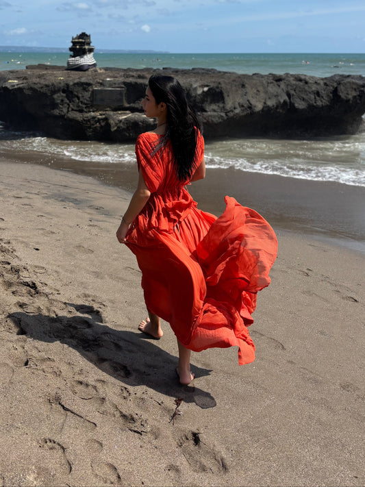 Woman in a red dress standing on a beach with ocean and rock formation in the background. RUH Agni Silk Set in orange rust – luxury slow fashion coord crafted by artisans in Chandigarh, blending Indian heritage with modern design.