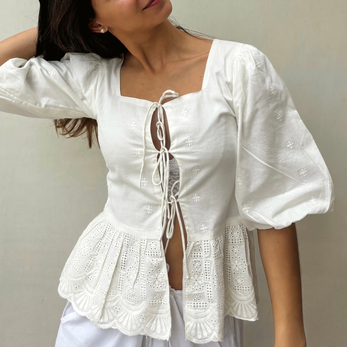 White lace-up top worn by a person against a neutral background. RUH Muse Blouse in pure cotton Chikankari embroidery – handmade white blouse with puff sleeves and bow ties.
