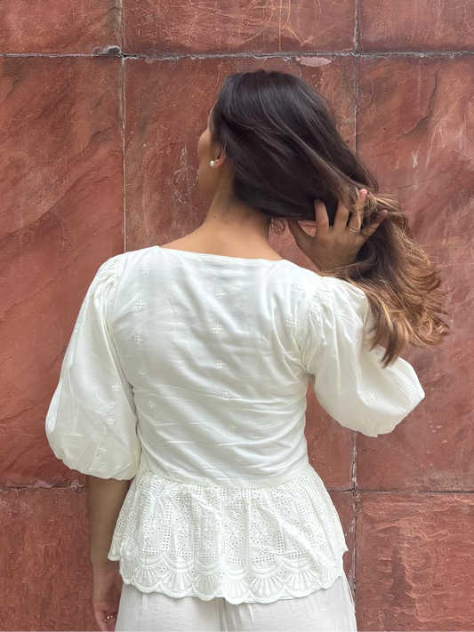 Woman in a white blouse standing against a red brick wall. Detail of RUH Muse Blouse bow ties and puff sleeves, inspired by Indian heritage reimagined for global wardrobes.