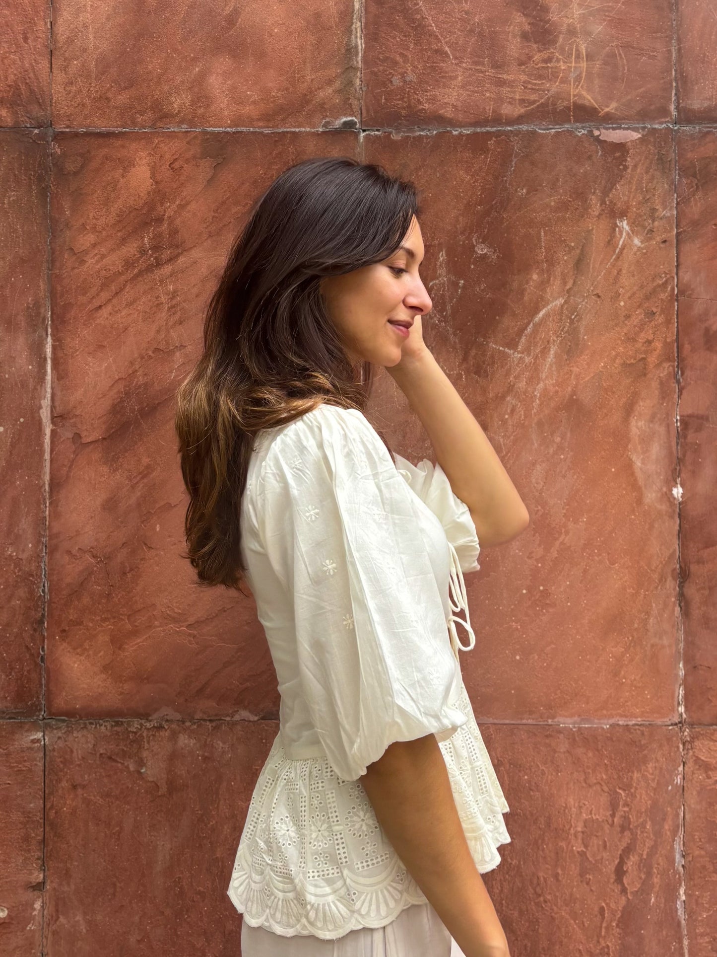 Woman in a white outfit standing against a textured brown wall. Detail of RUH Muse Blouse bow ties and puff sleeves, inspired by Indian heritage reimagined for global wardrobes.