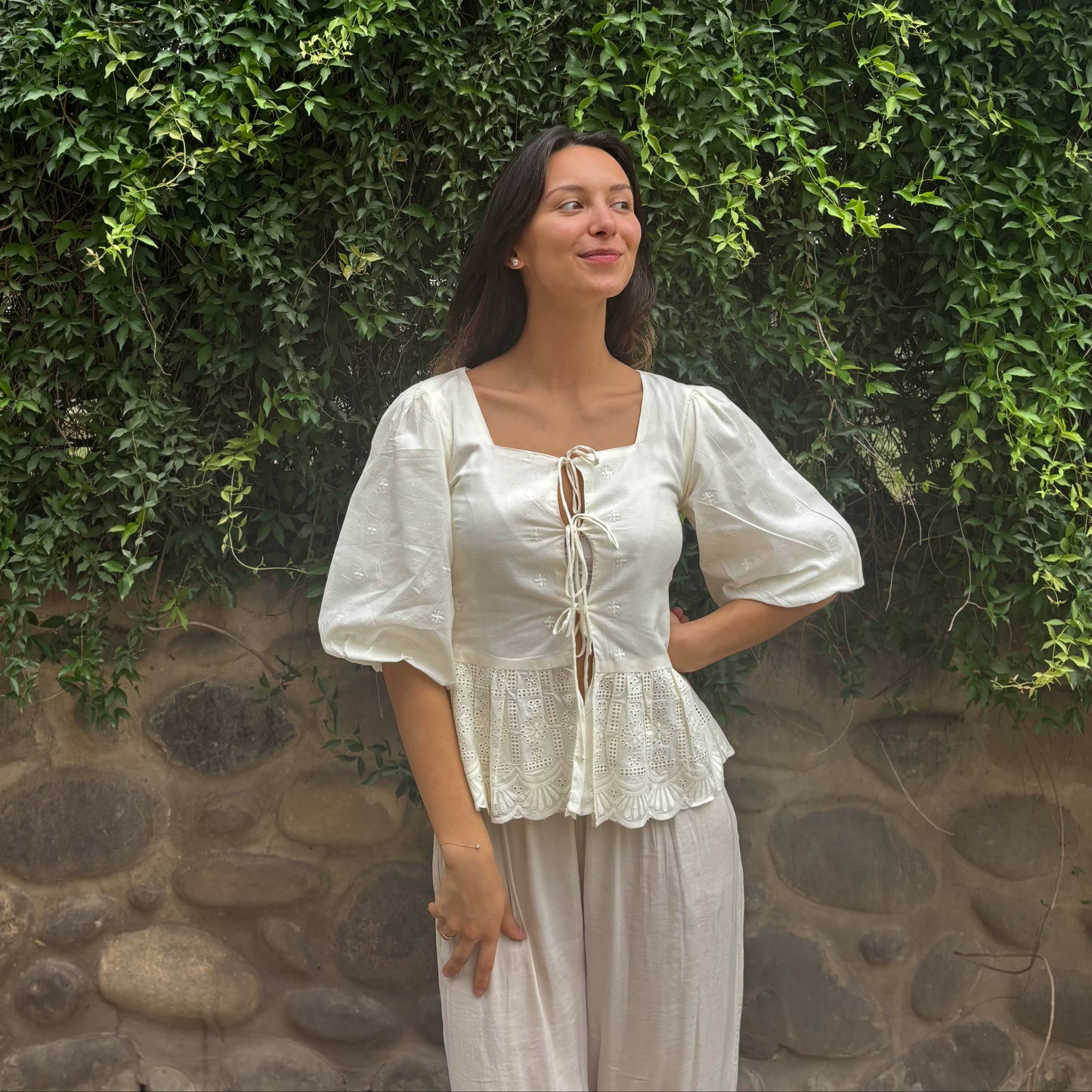 Woman in a white outfit standing against a stone wall with greenery. RUH Muse Blouse in pure cotton Chikankari embroidery – handmade white blouse with puff sleeves and bow ties.
