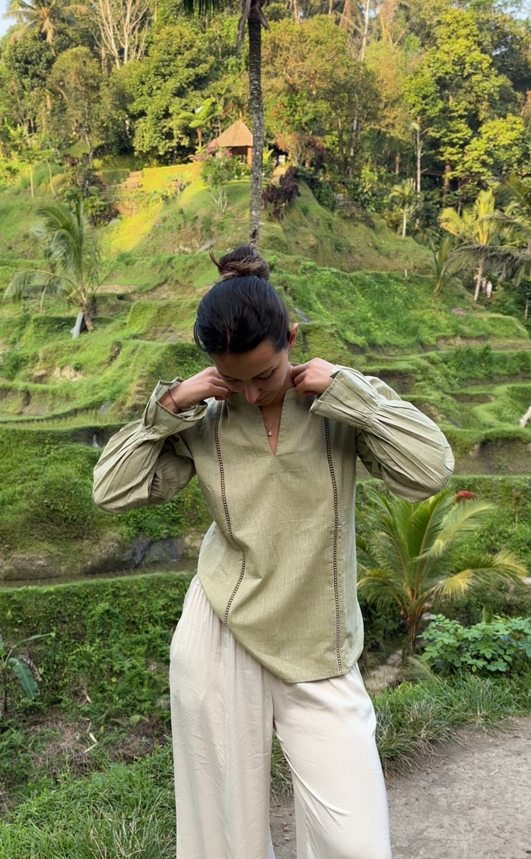 Person adjusting their shirt in a lush green outdoor setting with terraced fields and trees.