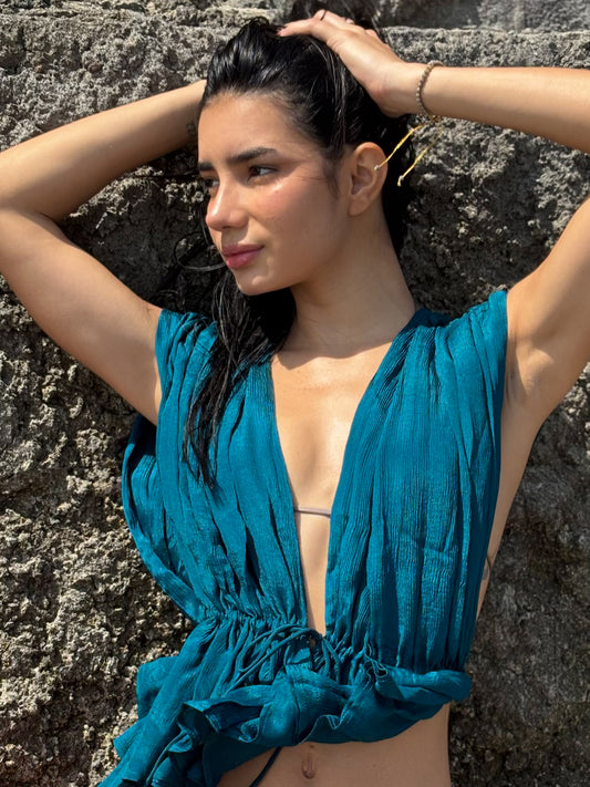 Woman in a teal top against a stone wall. Silk chiffon top with a front opening, low neck, adjustable ties. 