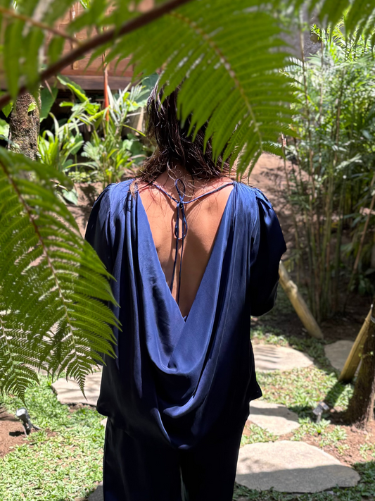 Person wearing a blue outfit walking through a garden with greenery and stones. Draped cowl-back satin silk top from RUH Aurora Set, slow fashion luxury with cultural fusion aesthetics. Low-rise wide-leg satin silk pants from RUH Aurora Set, handmade by artisans in India with premium finish.