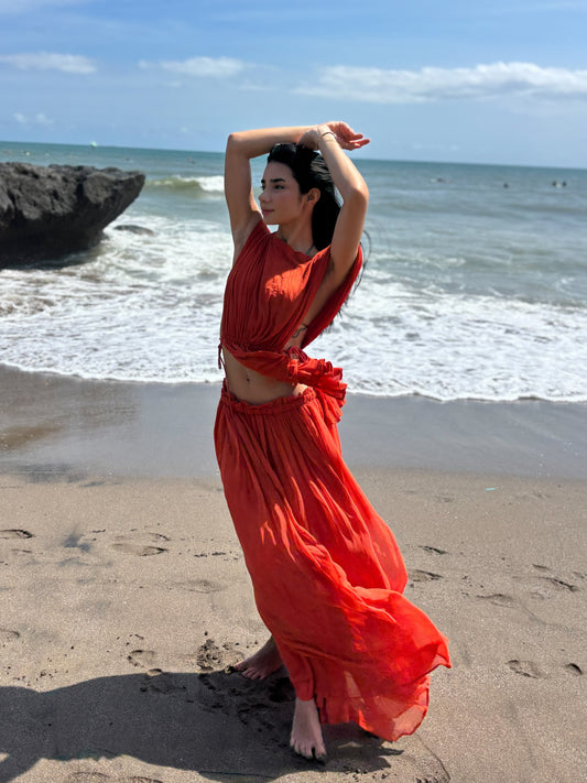 Woman in a red dress standing on a beach with ocean and sky in the background. RUH Agni Silk Set in orange rust – luxury slow fashion coord crafted by artisans in Chandigarh, blending Indian heritage with modern design.